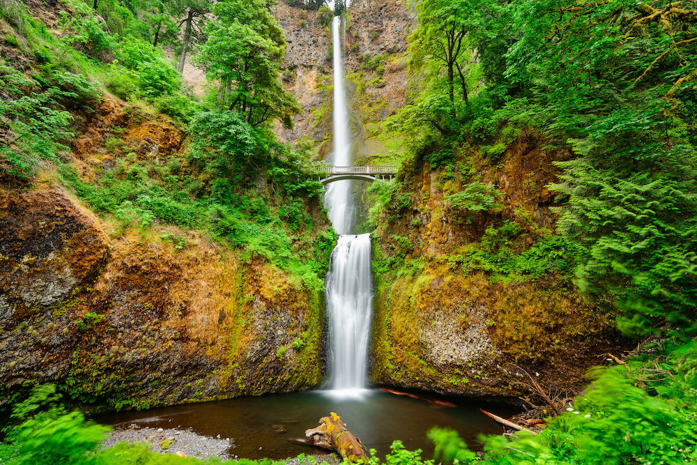 Multnomah Falls, Oregon, USA. Water cascading down a mountain, with a footbridge in view and greenery faming the falls.